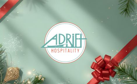 the adrift hospitality logo in blue and red on a mint green back ground with shiny red ribbon over the corners, pine greenery, gold ornament and sparkle accents. 