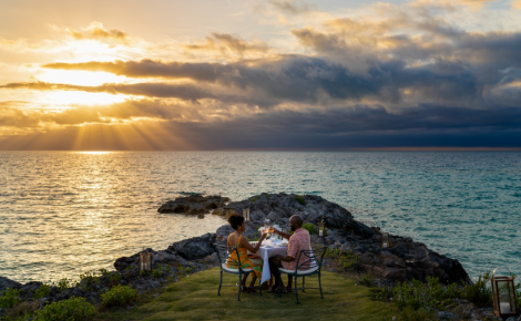 Couple enjoying a private oceanfront dinner on a grassy cliff at sunset, overlooking the turquoise waters of Bermuda.