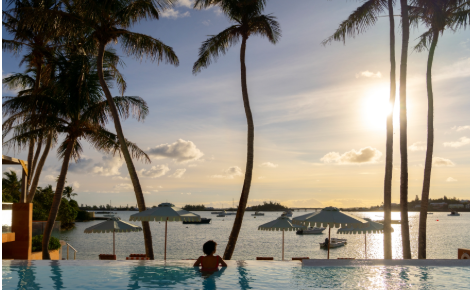 Guest relaxing in an infinity pool overlooking the bay at sunset with palm trees and boats in Bermuda.