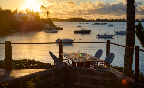 Oceanfront dining table overlooking a calm Bermuda harbor at sunset with boats anchored in the water.