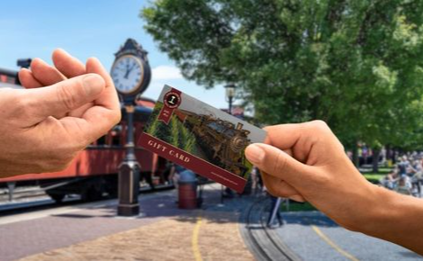 
A close-up shot shows one person’s hand passing a Strasburg Rail Road gift card to another person at an outdoor train station. The background features a vintage steam train, a classic station clock, and lush green trees under a bright blue sky.