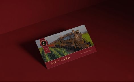 Image offers a stylized mock-up of a single Strasburg Rail Road gift card. The card is angled diagonally, resting on a flat surface within a minimalist, monochromatic setting. Both the surface and the backdrop are a solid, deep maroon-red (hex #670c12), creating a sophisticated and dramatic contrast