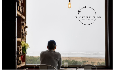 man in a gray long sleeve shirt and blue baseball cap with back to the camera facing large ocean front window with shelves of firewood to his left.