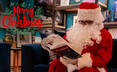 Image of Santa Claus reading a book on flight in the Ames Library at The Ameswell Hotel, with the words "Merry Christmas"