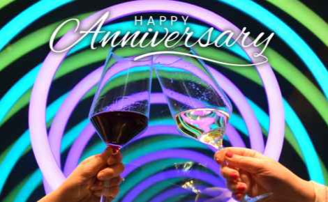 An image of two wine glasses being cheers'd in front of artwork at The Ameswell Hotel, with the words "Happy Anniversary"