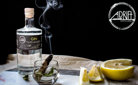 A bottle of Adrift distillers Gin in the foreground. There's a smoking bundle of herbs, and several cut lemons. The adrift distillers logo is in the top right corner.