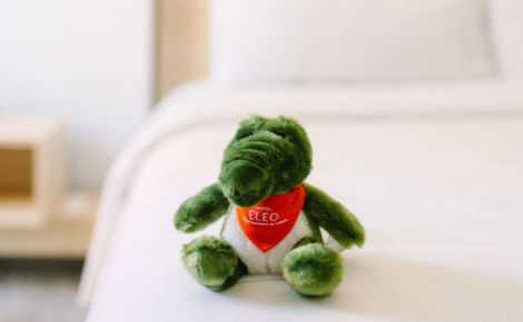 A plush alligator sits on the bed. The alligator has an orange bandana on that says Hotel ELEO. 