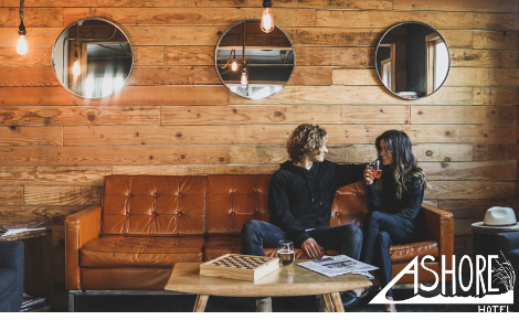 A wooden wall with three round mirrors is behind a leather couch. A couple sits laughing. The ashore logo is in the bottom right corner.