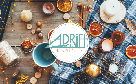 Adrift hospitality logo in the middle. In the background, a random holiday-ish spread of lights, ornaments, pinecones, string, oranges and candles lay atop a wooden table and blue plaid tablecloth.