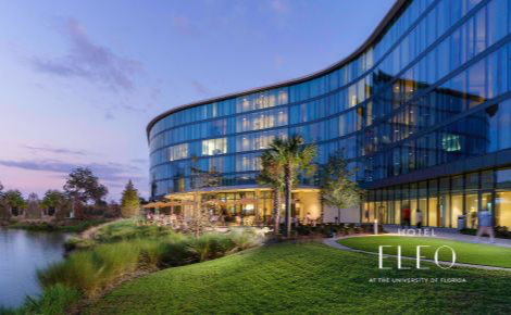The image is of the hotel at sunset with the lights glowing, people walking outside and a small view of the lake that the restaurant and hotel rooms overlook.