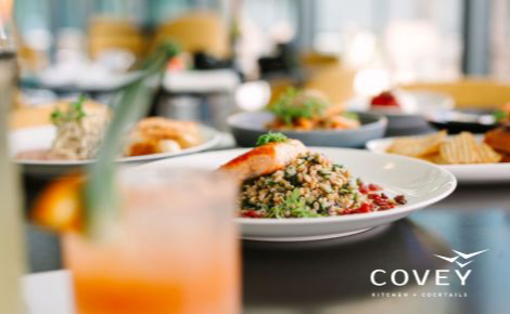 The table is full of mouth-watering dishes. This photo is of Covey Kitchen + Cocktails. A cocktail is close to the lens of the camera and is blurred but the orange of the drink glows. 
